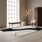 A modern abstract area rug with a black and off-white design is styled in a minimalist, warm-toned living room with a wooden floor, a leather chair, and a large window.