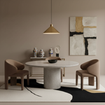 A modern dining room setup featuring a round concrete pedestal table, three upholstered armchairs in taupe, a black bowl centerpiece, and a warm brass pendant light. The furniture is arranged on a black and white abstract-patterned rug against a beige wall with a large abstract canvas print.