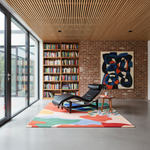 A modern black leather chaise lounge chair sits on a colorful abstract-patterned area rug in a stylishly decorated room with a brick wall, wooden slat ceiling, bookshelves, and a large abstract painting.