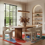 A modern dining room scene featuring a round wooden dining table surrounded by six cream-colored upholstered chairs, set on a colorful abstract-patterned rug. The room has warm beige walls, a built-in arched shelf with decorative ceramic vases, and a large window with sheer white curtains.