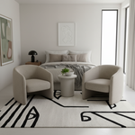 A modern living room setup featuring two beige upholstered armchairs facing a small round side table with a plant, set on a black and white geometric patterned area rug. In the background is a neatly made bed with neutral-toned bedding and minimalist wall art.