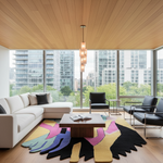 A modern living room with a large white sectional sofa, black leather armchairs, a low wooden coffee table, and a bold multicolor abstract rug. The room has a light wood slatted ceiling, large floor-to-ceiling windows showing a city skyline, and a central pendant light.