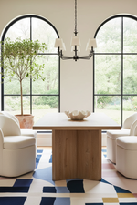 A modern dining room scene featuring a rectangular light oak wood table with a thick block pedestal base, surrounded by cream upholstered chairs. The room is styled with a geometric patterned rug, a black chandelier, and large arched windows looking out to a green garden.