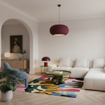 A modern living room featuring a prominent ribbed pleated pendant light in a deep burgundy/plum color hanging above a white sectional sofa. The room is styled with a colorful abstract patterned rug, a green coffee table, a blue armchair, and light wood flooring.