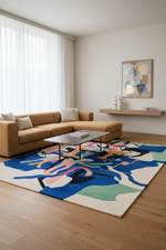 A modern living room featuring a large abstract area rug with blue, pink, green, and beige shapes on a light wood floor. A tan sectional sofa and a glass-top coffee table sit on the rug, with a small wall shelf and framed abstract art on the wall in the background.