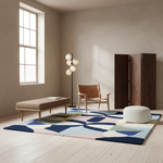 A styled interior room with a large abstract area rug in navy, blue, pink, beige, and green tones, a wooden bench, a leather chair, a floor lamp, a white pouf, and a dark wood cabinet.