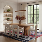 A modern dining room scene featuring a round wooden dining table surrounded by six white upholstered chairs with curved backs, set on a colorful geometric area rug. A large vase with autumn-colored foliage sits on the table. The room has white walls with built-in shelving, a hanging linear light, and a large black-framed window looking out onto green trees.
