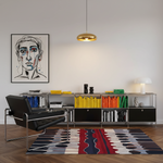 A modern living room scene with a black leather lounge chair, a gold pendant light, a white wall with a framed abstract portrait, and a low metal bookcase filled with colorful books and decor. A patterned area rug is visible on the wood floor.