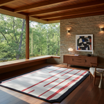 A large rectangular area rug with a cream base, bold red linear stripes, and a dark navy border, styled in a modern mid-century modern living room with a wood floor, stone wall, and large windows overlooking a green forest.