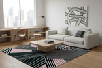A modern living room featuring a large black, white, and pink geometric patterned area rug on a light wood floor. The room includes a white sofa with accent pillows, a light wood coffee table, a built-in wooden desk and chair by a large window, and a black and white abstract canvas print on the wall.
