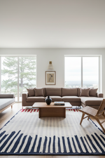 A modern living room featuring a large brown sectional sofa, a striped navy and cream area rug, a wooden coffee table, and a woven accent chair, with large windows in the background.