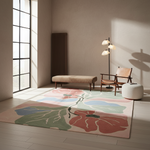 A large area rug with a stylized floral pattern in muted pink, green, and terracotta tones, styled in a modern minimalist living room with a wooden chair, floor lamp, and white pouf.