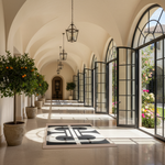 A long, elegant corridor with arched ceilings, black-framed glass doors opening to a garden, hanging lantern lights, potted orange trees, and a black-and-white geometric rug on the floor.