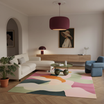 A styled living room interior with a cream sectional sofa, blue accent chair, round green coffee table, abstract multicolor area rug, wood console, and a large burgundy pendant light over a herringbone wood floor.