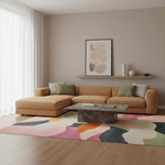 A modern living room featuring a tan sectional sofa with green and gray accent pillows, a marble coffee table, and a large abstract-patterned rug in pink, teal, orange, and cream. The room has beige walls, a floating shelf with decorative items, and a window with sheer white curtains.