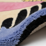 Close-up detail of a textured area rug showing a pattern of black abstract shapes on a cream background, bordered by a dusty blue tufted edge, with pink and yellow sections visible in the background.