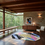 A modern living room interior with hardwood floors, a stone wall, large windows, a wooden bench, sideboard, chair, and a colorful abstract hexagonal rug on the floor.