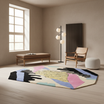 A modern abstract area rug with black, pink, blue, yellow, and beige sections is styled in a minimalist, warm-toned living room with a wooden bench, armchair, floor lamp, and white pouf.