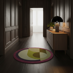 A round area rug with an abstract geometric pattern in olive green, mustard yellow, cream, and magenta, placed on a dark wood hallway floor next to a wooden sideboard with a lamp.