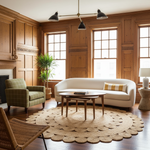 A styled living room with warm wood-paneled walls, a cream curved sofa, a green plaid armchair, a round natural jute rug, a wooden coffee table, and large multi-pane windows.
