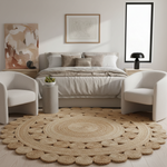 A styled bedroom scene featuring a large round natural jute rug on a light wood floor, with a bed, two cream armchairs, side tables, and framed wall art in a bright neutral interior.