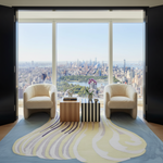 A luxury modern living room setup with two cream bouclé armchairs facing a low wooden and striped coffee table, all arranged on a large abstract blue, yellow, and white area rug. The room is situated in a high-rise apartment with floor-to-ceiling windows looking out onto a panoramic view of the New York City skyline and Central Park.