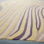 A close-up of a textured area rug with layered, wavy organic stripes in pale yellow, cream, taupe, and muted purple, with a soft blue-gray border visible at the bottom.