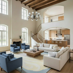 A modern, high-ceilinged living room with a spiral staircase, featuring a cream sectional sofa, two blue armchairs, a wooden coffee table, and a large abstract area rug. The room has high windows, exposed wood ceiling beams, and an open kitchen in the background.