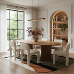 A modern dining room featuring a light wood oval table with six cream upholstered chairs, set on a geometric patterned rug. The room has warm white walls, a built-in arched shelf with warm lighting, and a large window looking out onto green trees.