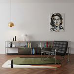 A modern black leather tufted lounge chair with a chrome frame sits on a green and cream abstract area rug in a minimalist living room. A low metal shelving unit with books and decor is against a white wall, featuring a black-and-white portrait painting above.