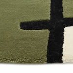 Close-up of a textured area rug with a geometric pattern in olive green, black, and off-white, showing the pile texture and fringe edge.