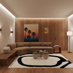 A modern living room featuring a curved tan sectional sofa, a large black and white abstract area rug, a glass-top coffee table with stone-like stools, and a warm wood-paneled wall with a sunset palm tree painting.
