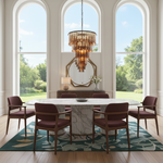 A luxury dining room featuring a round white marble dining table surrounded by six modern armchairs with dark burgundy upholstery and wood frames. The furniture is arranged on a teal and beige botanical patterned area rug. Above the table hangs a multi-tiered gold-toned crystal chandelier. The room is set in a bright, elegant dining room with tall arched windows looking out onto a green lawn, a cream console table, and a large hexagonal mirror.