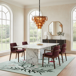 A modern dining room featuring a white marble rectangular table with a marble pedestal base, surrounded by four dark wood chairs with burgundy upholstered seats. A light green floral area rug covers the floor, and a copper-toned glass chandelier hangs from the ceiling. Large arched windows with white frames look out to a green garden.