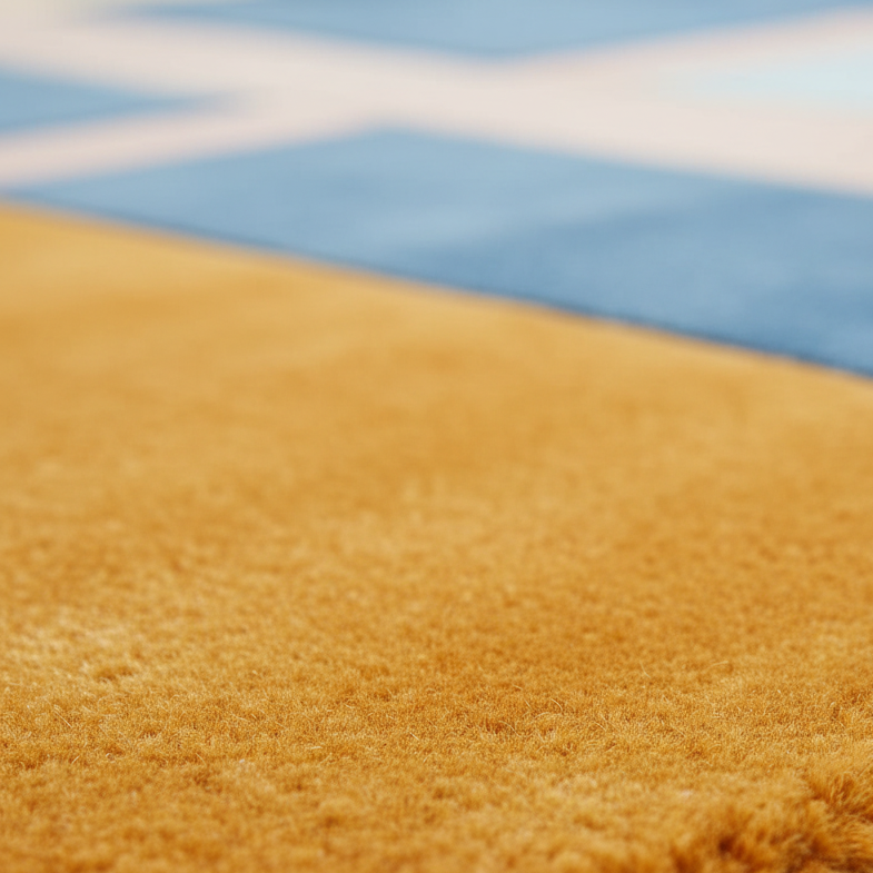 Close-up of a plush, high-pile mustard yellow or golden-brown rug, with a soft-focus background showing a blue and white patterned rug.