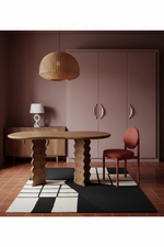 A styled interior scene with a round wooden dining table, sculptural wavy legs, a reddish-brown chair, a woven pendant light, a geometric black-and-white rug, and a muted pink wardrobe in the background.