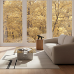 A modern living room with a light beige sofa, a glass-top coffee table, and a large white rug, set against floor-to-ceiling windows showing a bright golden autumn forest view.
