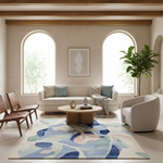 A modern living room featuring a large abstract area rug with blue, beige, and white floral patterns. The room is styled with a cream sofa, a round wooden coffee table, a white accent chair, and two wooden chairs, set against a warm beige wall with two arched windows.