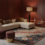 A modern living room scene featuring a large area rug with a floral and abstract pattern in muted blue, pink, cream, and gold tones. The rug is styled in a contemporary room with a cream sectional sofa, dark wood-paneled walls, a floor lamp, and decorative side tables.