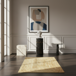 A styled interior scene with a framed abstract wall art print above a black marble pedestal, alongside a woven natural-fiber rug, a white marble cube, and a small black side table in a sunlit room.