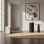 A styled interior scene with a framed abstract wall art print above a black marble pedestal with a white bust, alongside a small black side table and a round beige rug on dark wood flooring.