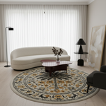 A round area rug with an intricate floral and palm tree pattern in beige, yellow, black, and gray, styled in a modern living room with a curved cream sofa, dark wood coffee table, and black floor lamps.