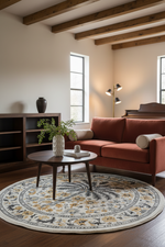 A styled living room featuring a rust-red sectional sofa, a round wooden coffee table, a large round patterned area rug, and a dark wood shelving unit against a warm beige wall with exposed wooden ceiling beams.