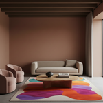 A modern living room scene with a beige sofa, two blush pink armchairs, a low wooden coffee table, and a large abstract area rug in orange, purple, teal, and pink on a light floor. The room has warm taupe-brown walls, dark wood ceiling beams, and a window with curtains.
