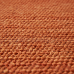 Close-up of a textured woven rug in a warm rust-orange color, showing the detailed loop pile and braided pattern.