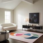 A modern living room featuring a large abstract area rug with overlapping pastel and cool-toned geometric shapes in orange, pink, cream, green, and blue on a pale blush background. The room includes a cream curved sofa, a black media console with a lamp and framed abstract wall art, and a window on the left.