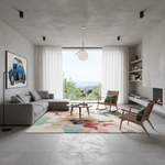 A modern living room with a large abstract multicolor area rug on a gray tiled floor, featuring a gray sectional sofa, wooden armchairs with green cushions, a round coffee table, and a large window with white curtains overlooking a distant landscape.