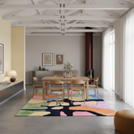 A large, colorful abstract-patterned area rug in a modern dining room setting. The rug features bold, organic shapes in pink, green, yellow, blue, and black. It is styled under a light wood dining table and chairs, with a gray concrete floor and a high ceiling with white beams.