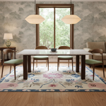 A styled dining room scene featuring a rectangular white marble-top dining table with dark wood legs, surrounded by four mid-century style chairs with light green cushions. The setup is on a large floral area rug with a light wood floor, set against a backdrop of patterned wallpaper, a window looking out to greenery, and two hanging pendant lights.