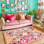 A vibrant floral area rug in a living room setting, featuring a hand-tufted wool design with pink and orange hues. The rug is situated in front of a cozy pink sofa adorned with colorful cushions. The walls are decorated with framed botanical art, creating a vintage and inviting atmosphere. Perfect for adding a touch of elegance to any living room area.
